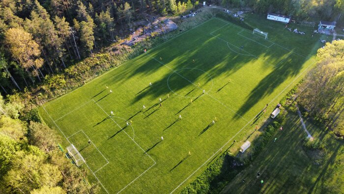 Fotbollsmatch på grönt fält med skuggor i solnedgång, sedd från ovan.