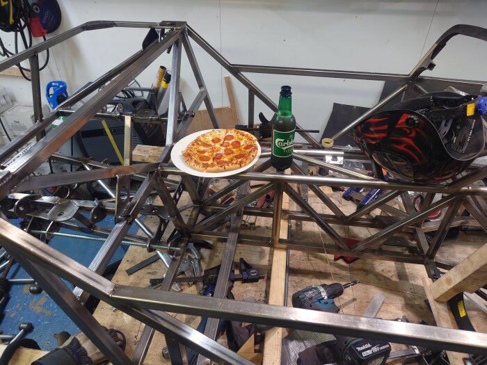 Metallramkonstruktion i verkstad med verktyg, hjälm, pepperonipizza och en flaska Carlsberg.