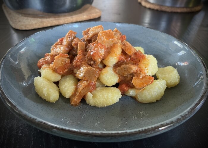 Gnocchi med röd pastasås och bitar av porchetta på en grå tallrik.