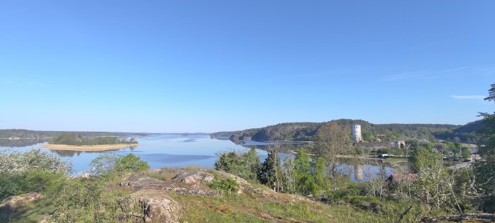 Vidsträckt utsikt över lugnt vatten med skog och en historisk tornbyggnad under en klarblå himmel.