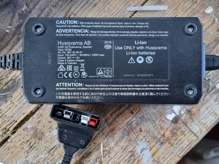 Grästrimmerbatteriladdare utan indikator-lampa med teknisk info och kontakt med fyra poler på träunderlag.