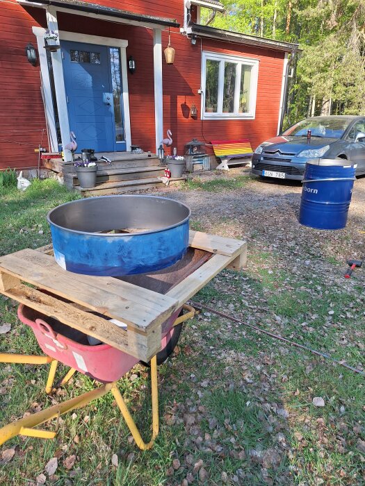 Ett omgjort blått fat monterat på en träkonstruktion som en skyddsring för plantering, framför röd husfasad.