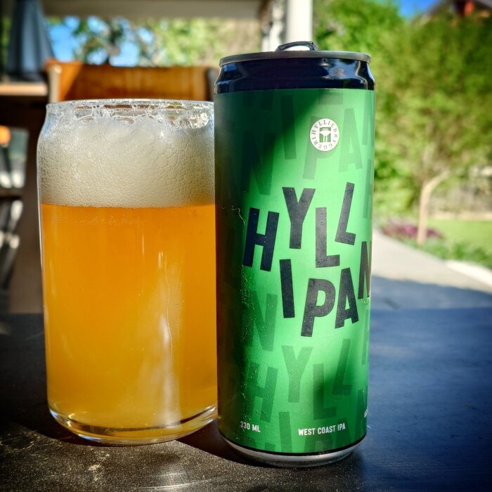 Ölburk märkt "HYLL IPA" bredvid ett glas med öl utomhus på ett bord.