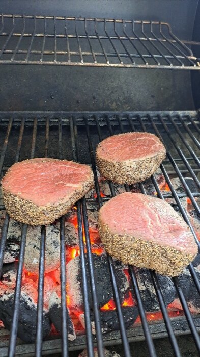 Tre stycken pepparkryddade biffar grillas över glödande kol på en grill.