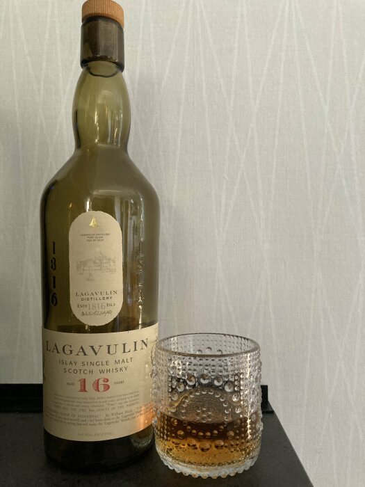 Flaska Lagavulin 16 år och ett whiskyglas med litet kvar, på bänk.