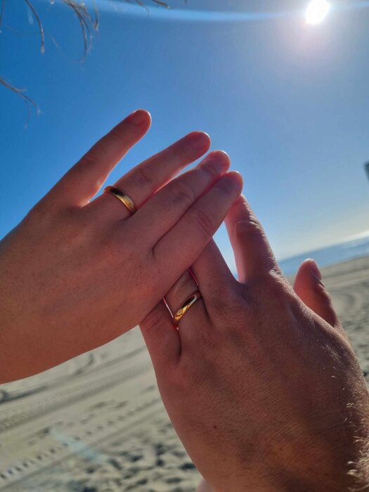 Två händer med vigselringar mot en blå himmel och solsken, antydande en frieri på stranden.
