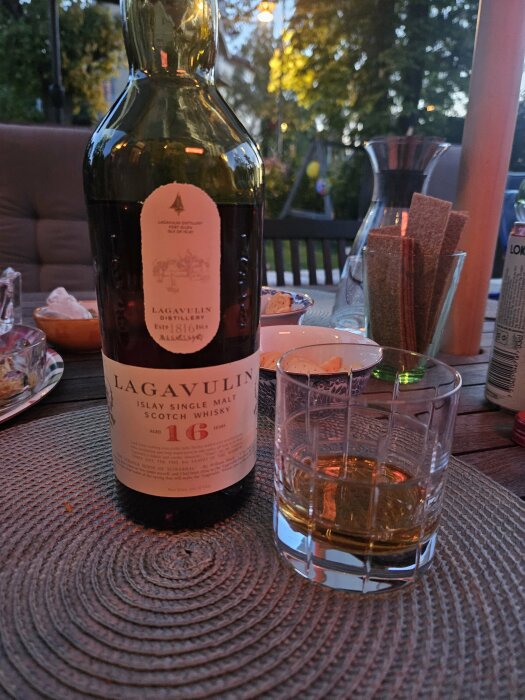 Flaska Lagavulin 16 år single malt scotch whisky med ett glas bredvid på ett utebord vid skymning.