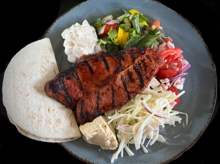 Grillad fläskkarré på tallrik med sallad, coleslaw, fetaost-tomatsallad, potatissallad och hummus.