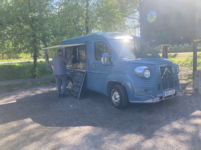 Blå food truck med öppet försäljningsfönster och en man som betjänar, parkerad utomhus på en solig dag.