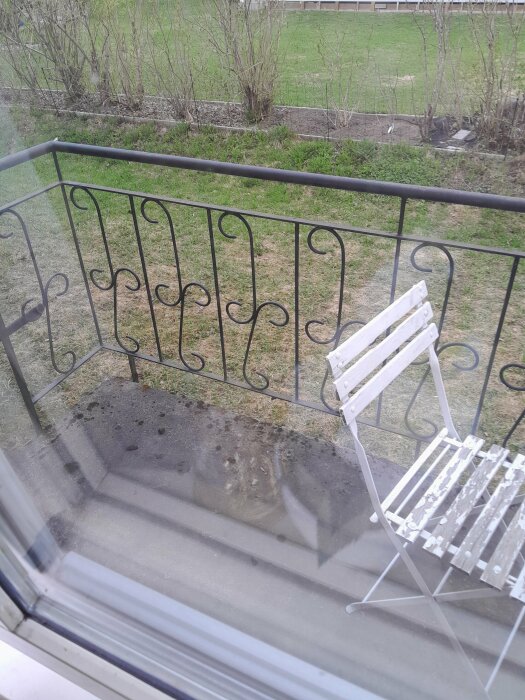 Svart smidesjärnsräcke på balkong med vita trädgårdsstolar, sedd genom fönster.