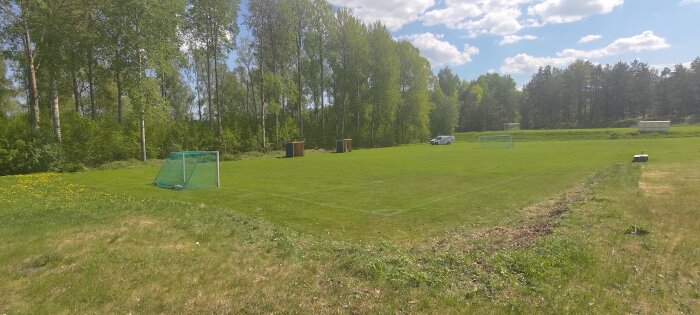 Välklippt 7-mannaplan för fotboll omgiven av nyklippt sly och träd, med fotbollsmål och skog i bakgrunden.