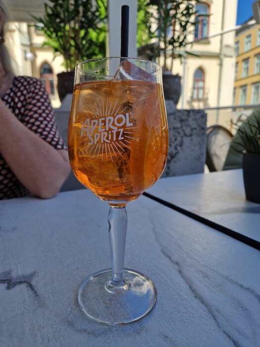 Aperol Spritz i glas på uteservering med isbitar och sugrör, i bakgrunden syns person och byggnader.