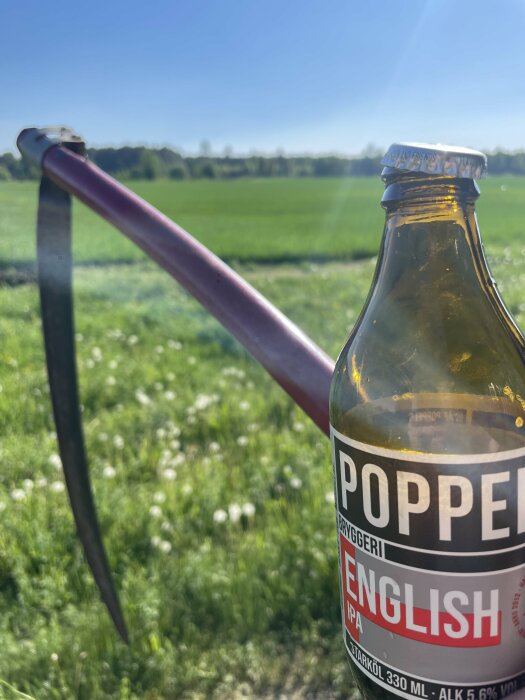 Flaska Poppels English IPA i förgrunden med suddig bakgrund av en grön äng och blå himmel.