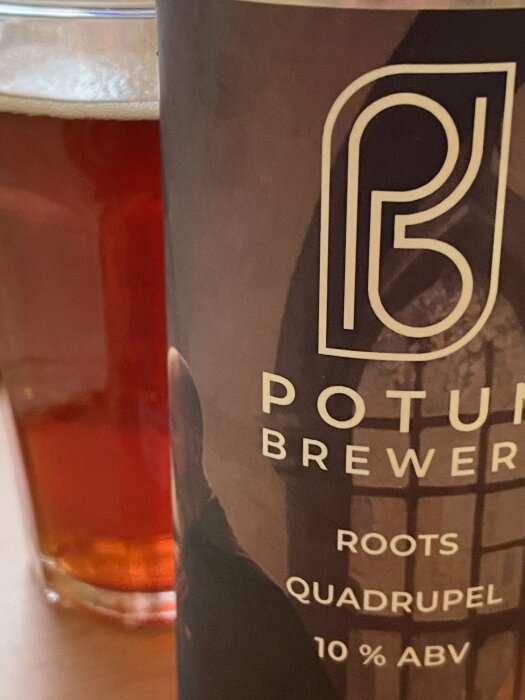 Ölglas delvis synligt med mörkbrunt öl bredvid en ölburk märkt "POTUS BREWERY ROOTS QUADRUPEL 10% ABV".