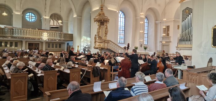 Publik i träbänkar lyssnar på symfoniorkester och kör framförande i kyrka.
