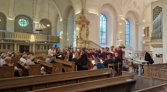 Kör och orkester framför musik i en kyrka med åhörare sittande i kyrkbänkar.