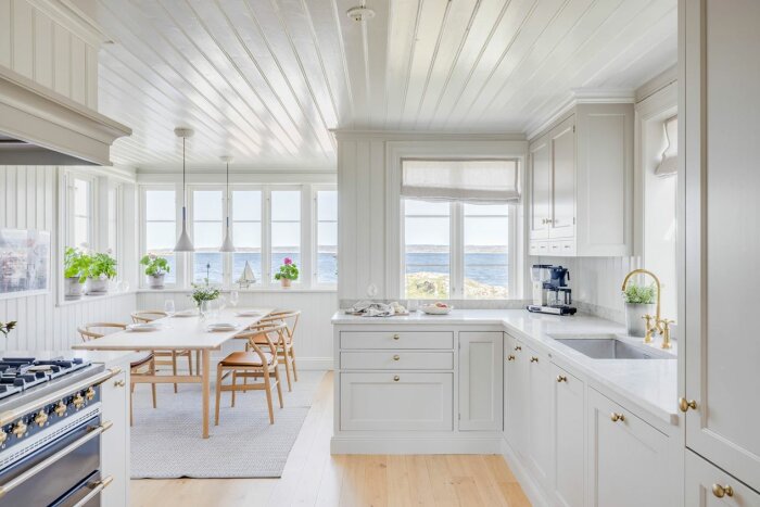 Ljust kök med vita skåp och träbord framför fönster med utsikt över havet.