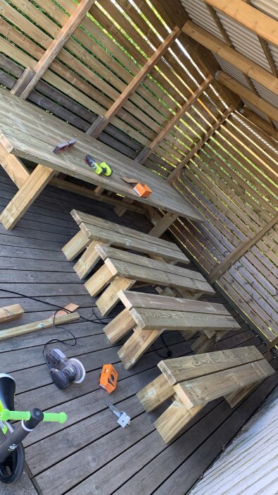 Hantverksprojekt av ett oavslutat trädgårdsbord med bänkar, verktyg och en barnsparkcykel på en träterrass.