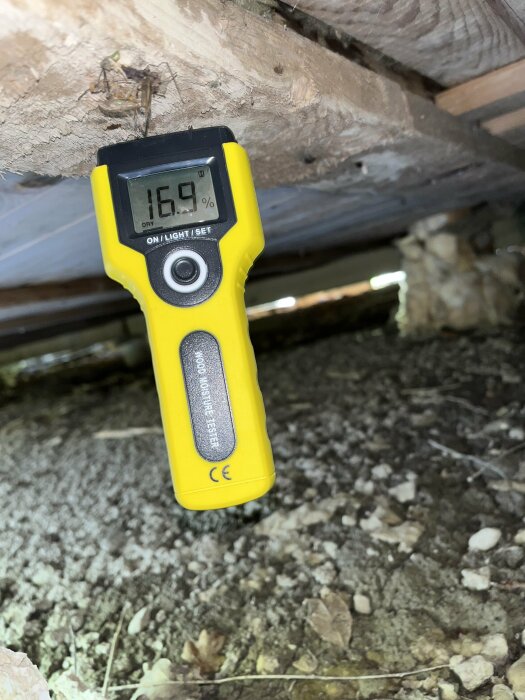 Fuktighetsmätare visar 16,9% på träbjälke i krypgrund under hus, mögel och pålagringar synliga.