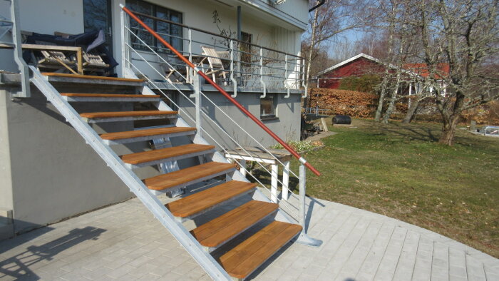 Renoverad trappa utomhus med trästeg och vit metallräcke mot en villa och trädgård i bakgrunden.