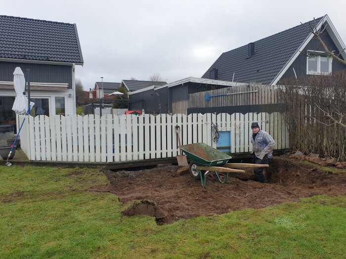 Person som gräver i trädgård för byggprojekt med skottkärra och staket i bakgrunden.