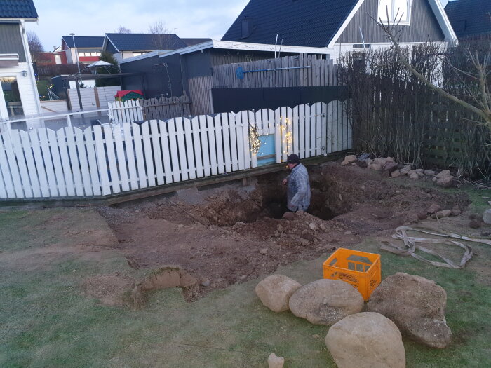 Person gräver en stor grop i en trädgård nära ett vitt staket, med gräsmatta och stenar framför.
