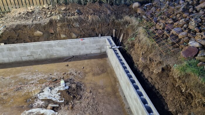Grundläggningsarbete med betongblock och armering i en grävd grundgrop.