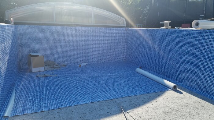 Halvfärdigt poolprojekt med blå mosaikplattor och byggmaterial på golvet i solljus.