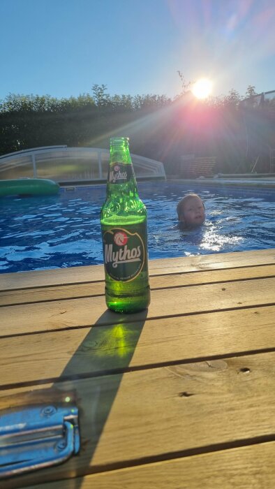 En Mythos-ölflaska på ett träbord med en simmande person i bakgrunden och solnedgång.