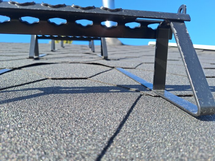 Närbild av takstege monterad på tak med asfaltssingel, skuggor från stege på solig dag.