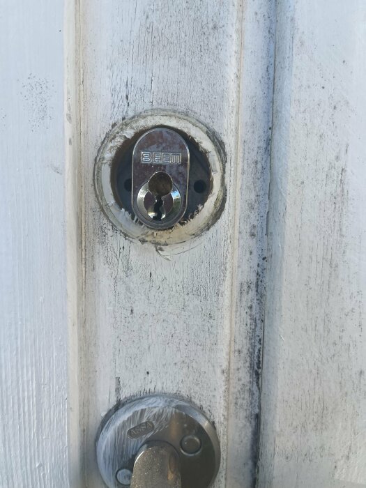 Närbild på ett vitmålat dörrlås med borrspår och en skruv avlägsnad, dörrens skicket är slitet.
