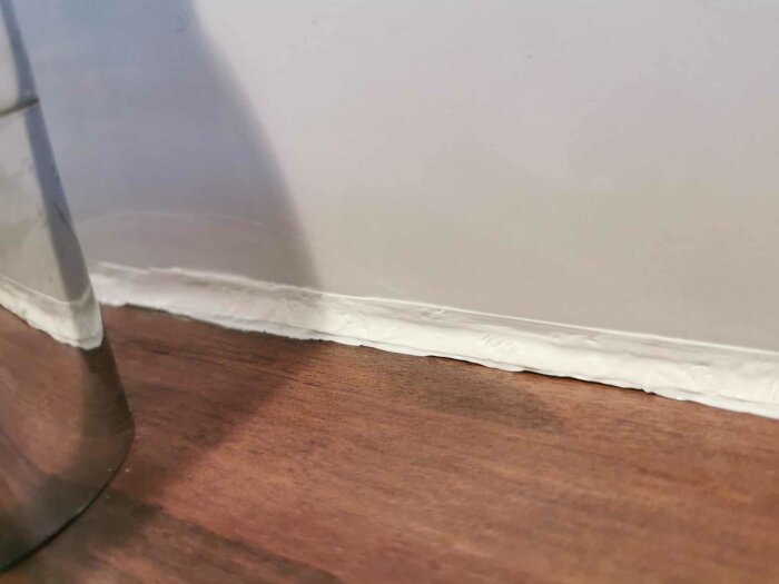 Närbild på en ojämnt applicerad vit fog längs skarven mellan en vit vägg och träfärgat golv.