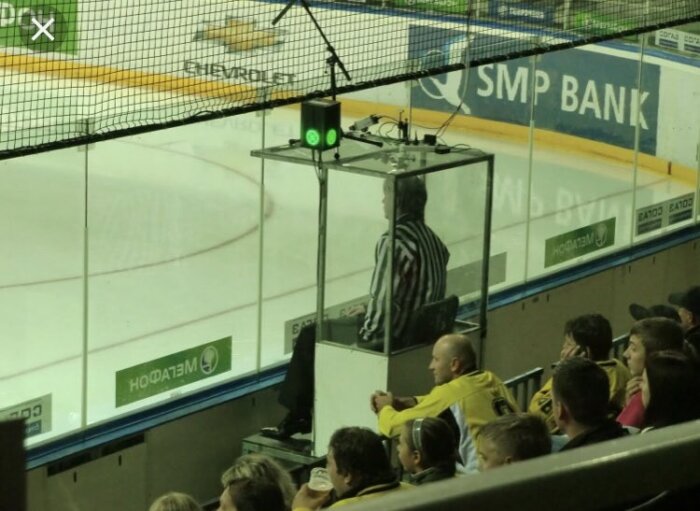 Måldomare i svart-vit randig tröja sitter i ett glasinburat upphöjt domartorn vid en hockeyrink.