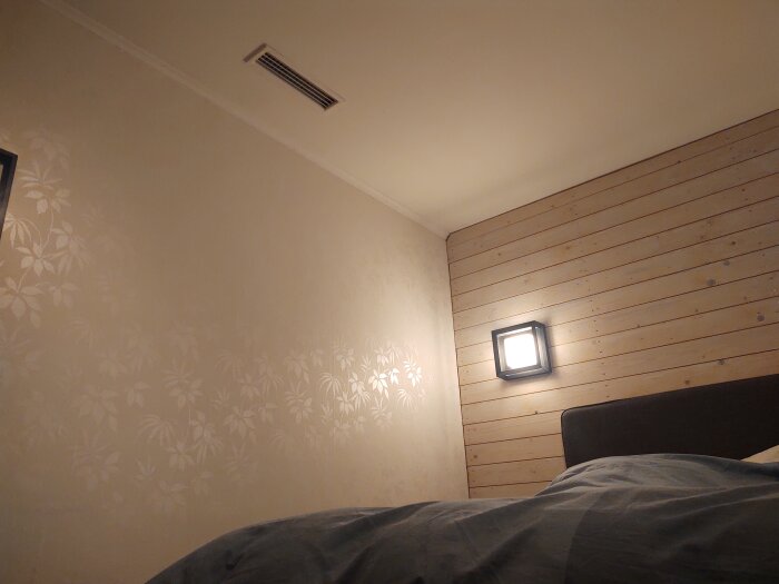 Ventil på vägg i sovrum med träpanel och blommönstrad tapet, medan en lampa är tänd.