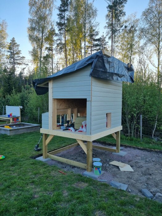 Upphöjt hönshus under konstruktion med isolerade väggar och påbörjade knutbräder, täckt av en svart presenning på taket.