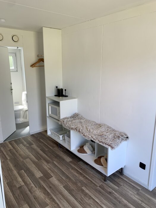 Renoverat köksutrymme med vit bänk, kuddar och vedgolv, dörr till badrummet synlig i bakgrunden.