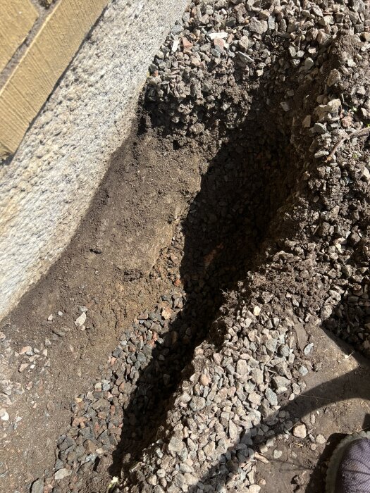 En närbild av en ojämn betongklack intill ett grävt dike fyllt med makadam och lerjord, vid förberedelser för dränering.