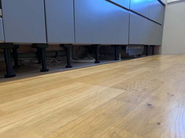 Nytt vinylklick golv i köket som möter bänkskåpens fötter, med rör och detaljer under skåpen synliga.