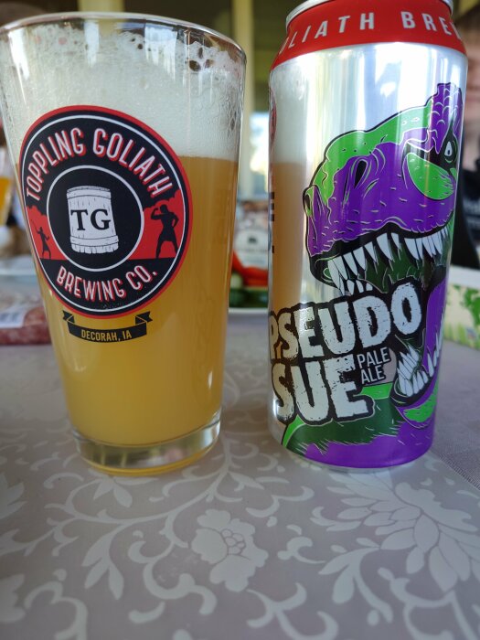 Ölglas med Toppling Goliath logotyp fyllt med grumlig dryck bredvid en Pseudo Sue Pale Ale burk.