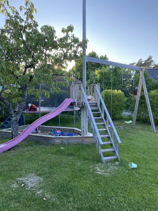 Hembyggd trälekplats med rutschkana, sandlåda och klätterställning i en trädgård.