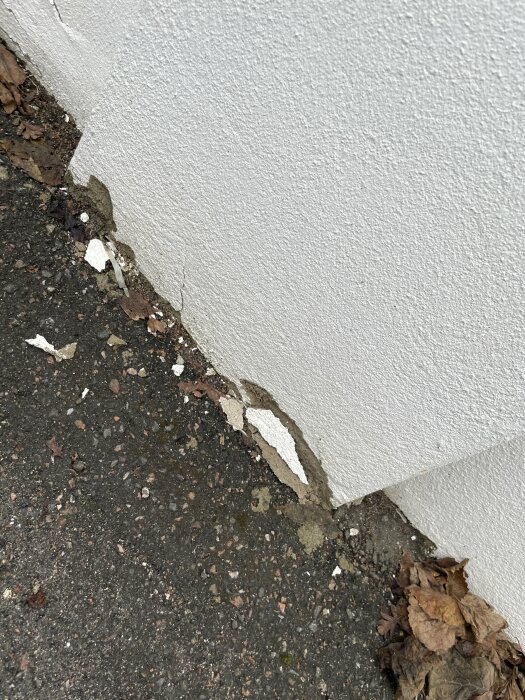 Skador på putsen vid botten av en vit stödmur där den möter asfalten, med synliga sprickor och avlagringar.