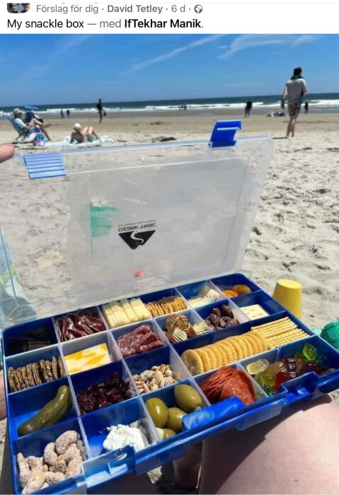 En sorteringslåda fylld med snacks som hålls upp framför en solig strand med badgäster.