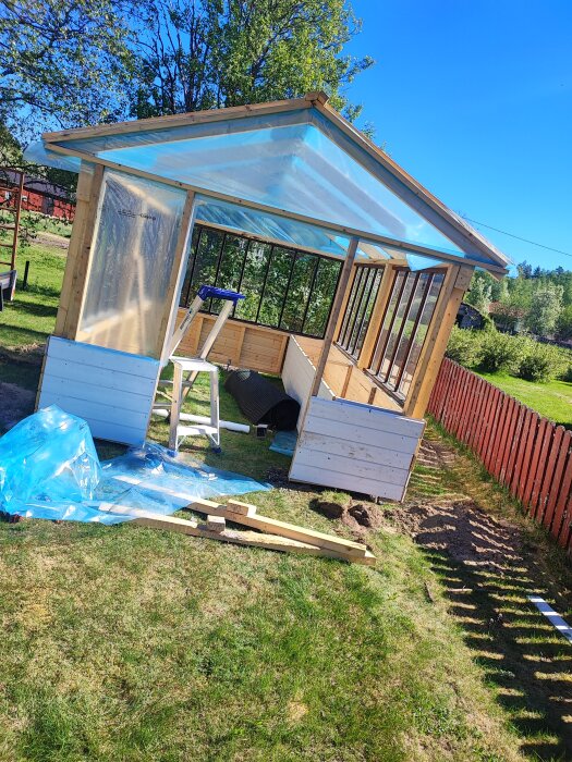 Pågående bygge av ett litet växthus med trästomme och genomskinligt tak, byggmaterial spridda på gräset.