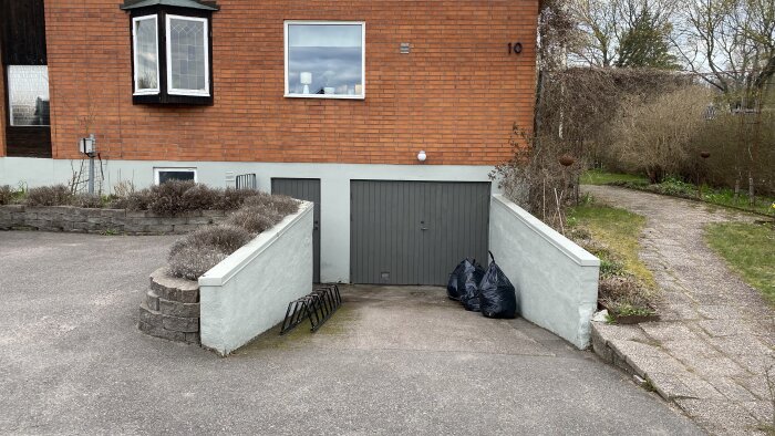 Garageinfart med höga betongstödmurar som leder ner till en grå garageport, omgiven av en trädgård och tegelhus.