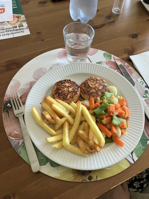 Tallrik med två laxburgare, pommes frites och grönsaksblandning på ett träbord.