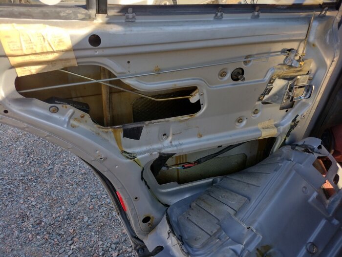 Insidan av en bilens bakdörr utan panel, synliga delar av kaross och mekanismer, tecken på rost.