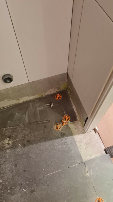 Oavslutat kakelarbete i ett hörn med synlig golvvärme och flera orange distanskors utspridda på golvet.