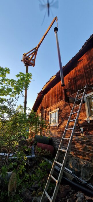 Skorsten lyfts av ett gammalt trähus med hjälp av mobilkran, stege lutad mot husväggen.