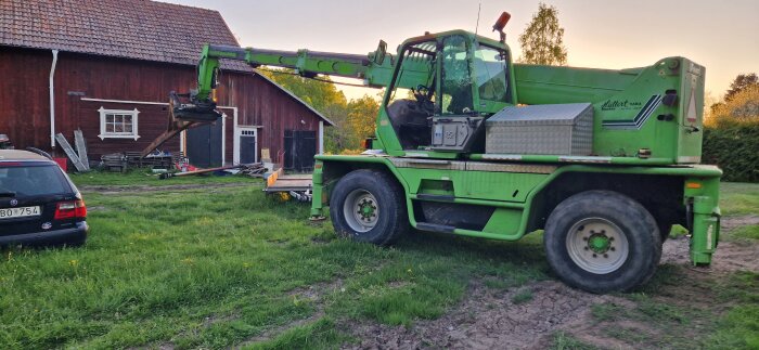 Grön kranbil lyfter en skorsten, bakom syns ett rött trähus och en parkerad bil.