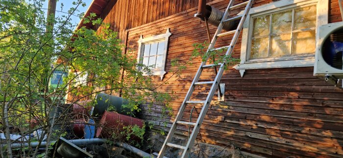 Stege lutad mot ett trähus med avtagen skorsten på taket och rosenbuskar i förgrunden, projektarbete pågår.